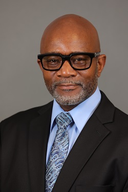 Man in a suit with glasses, smiling slightly against a gray background.