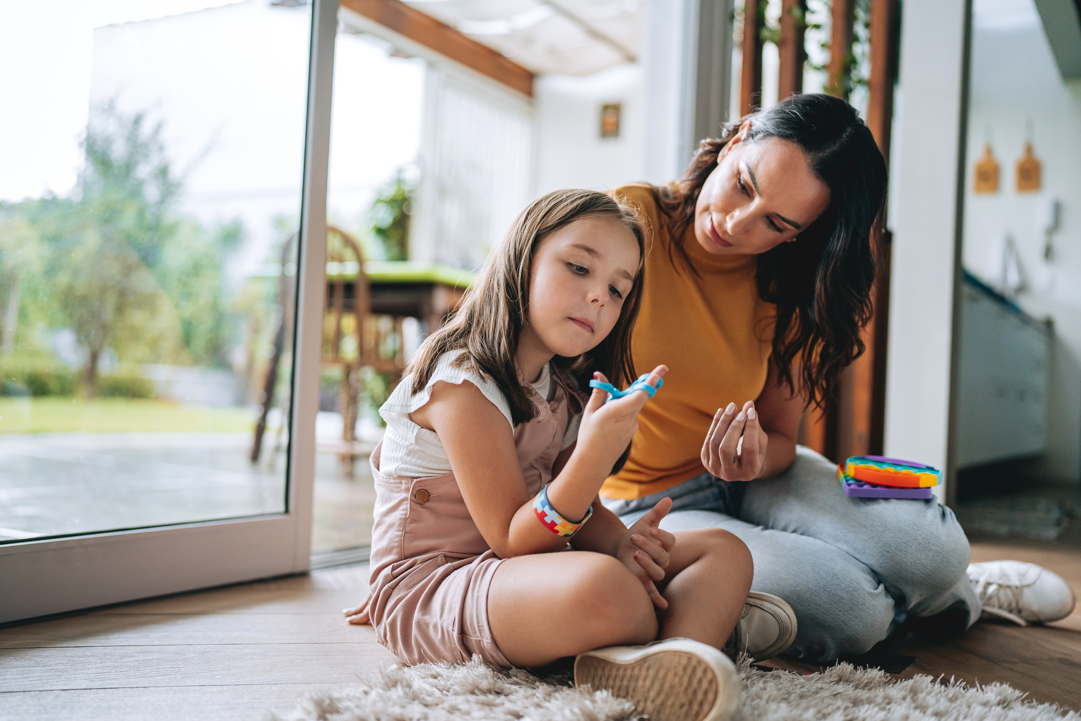Woman and young girl sitting on the floor painting together near a large window.
