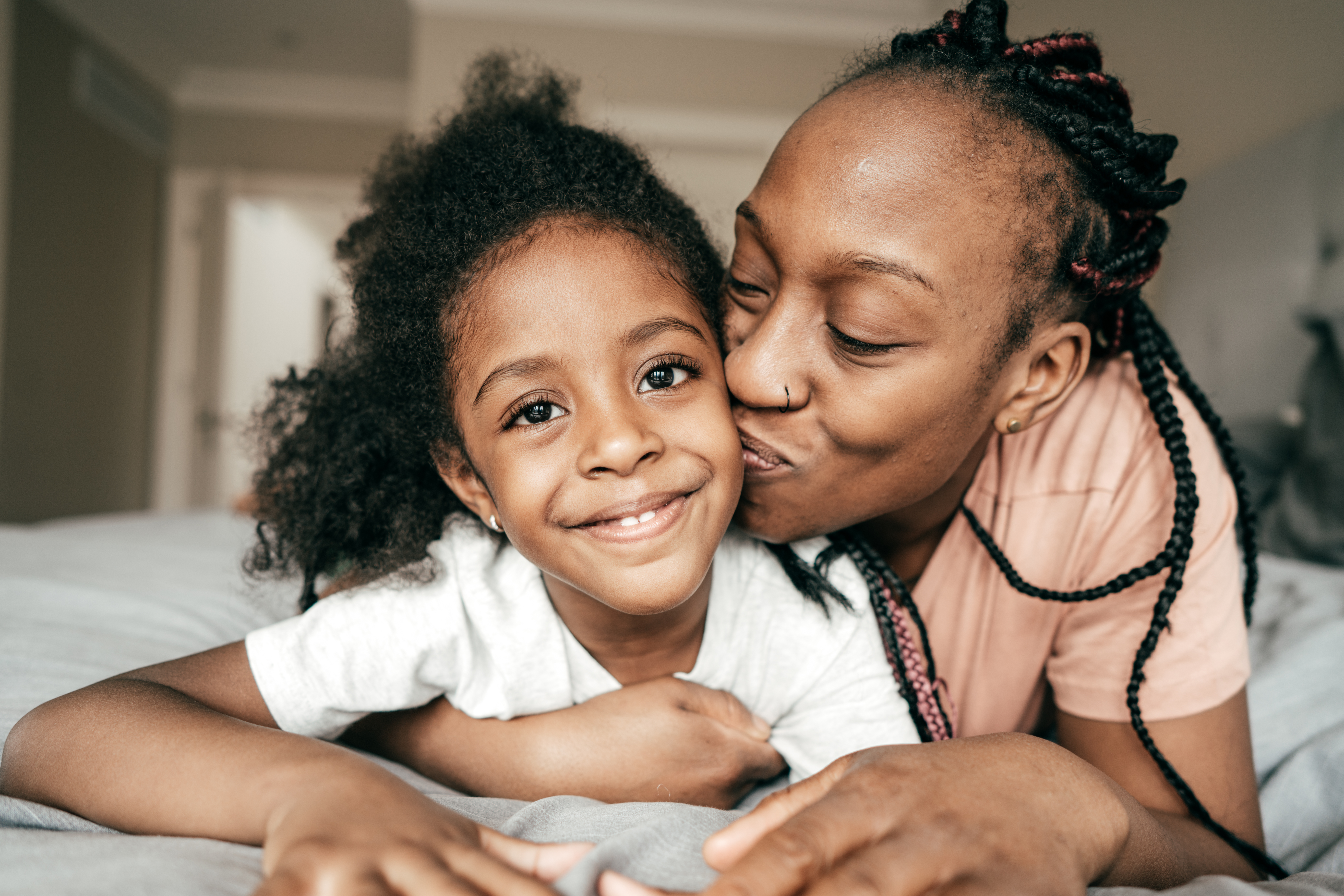 Woman kissing smiling child on the cheek while lying on a bed.