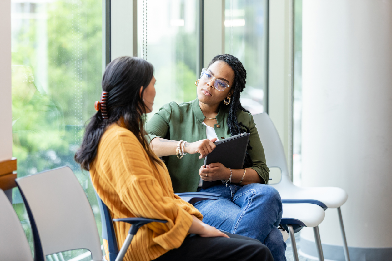 Two women sitting and talking by large windows in a bright, modern room.
