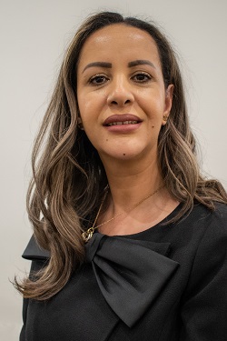Woman with long brown hair wearing a black top with a large bow, neutral background.