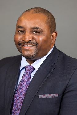 Man in a dark suit and purple tie smiling against a gray background.