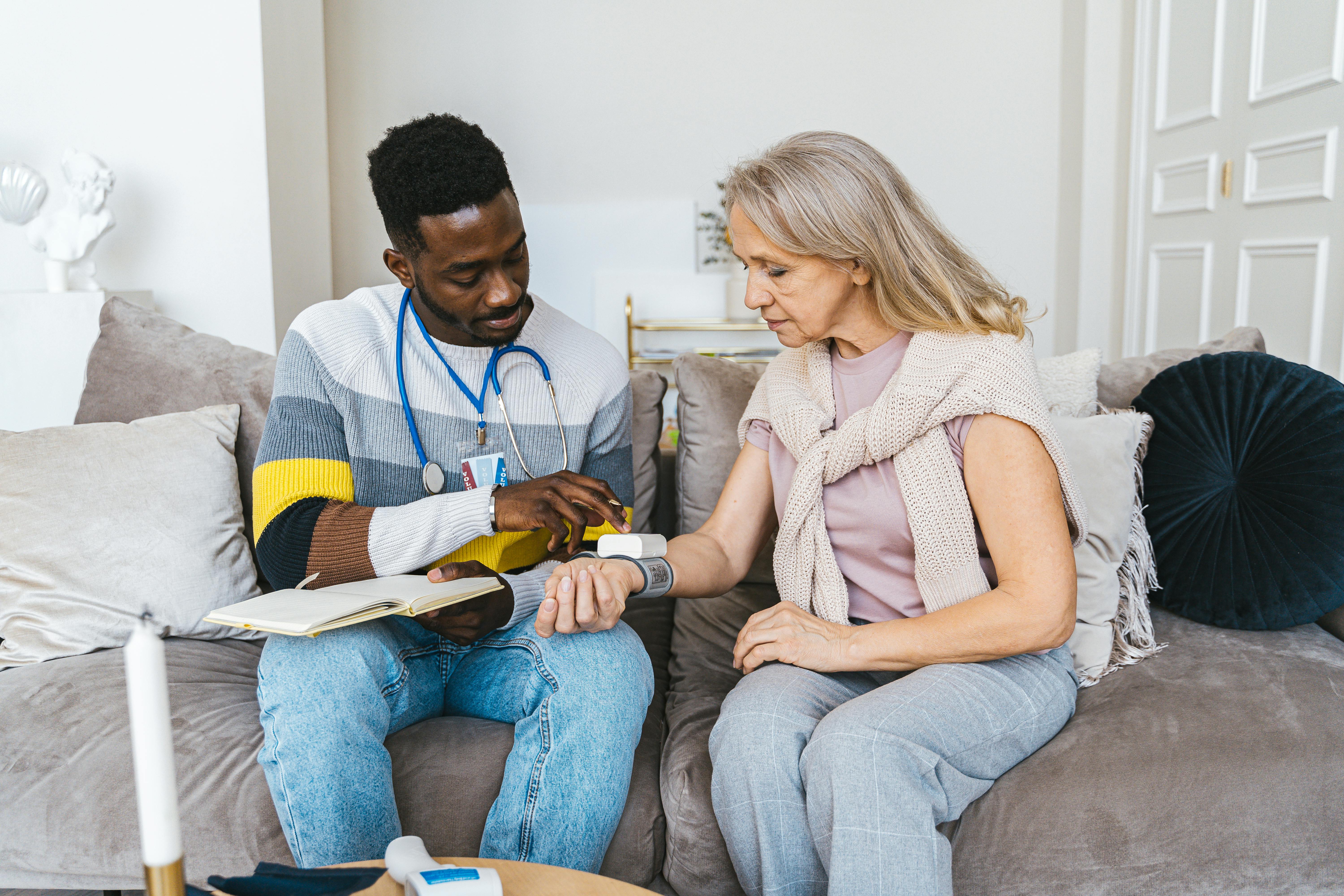 Doctor checking a woman’s blood pressure on a couch in a cozy living room.