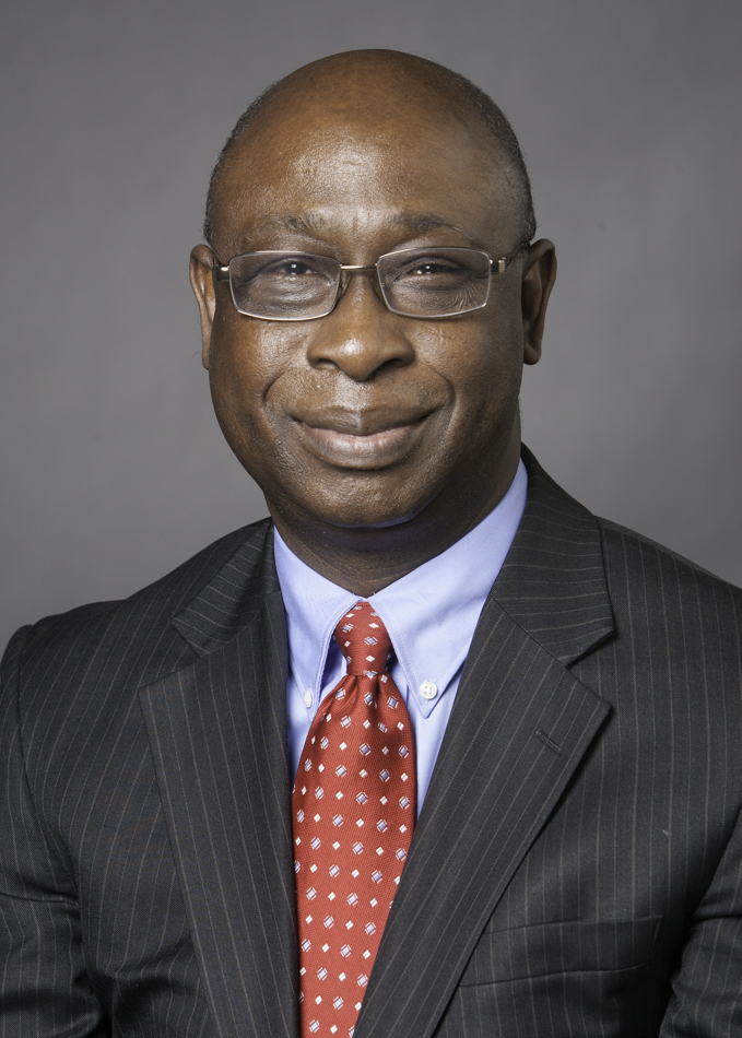 Man in a dark suit and red polka dot tie smiling against a gray background.