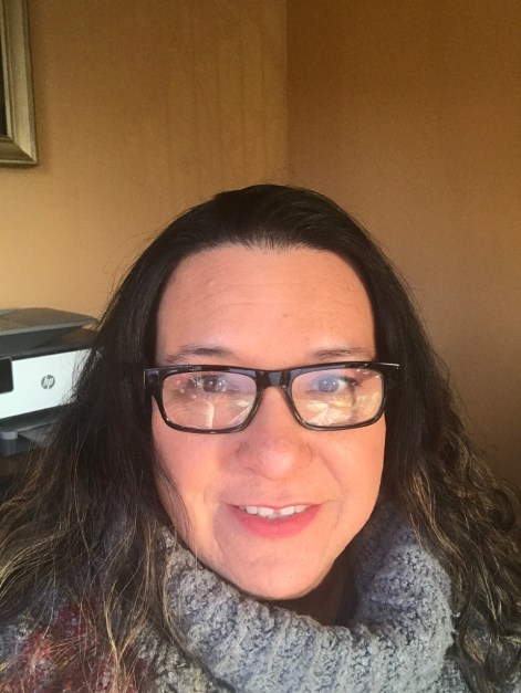 Woman with long dark hair and glasses smiling indoors, wearing a gray scarf.