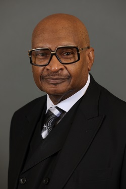 Man in a black suit and glasses against a plain gray background.