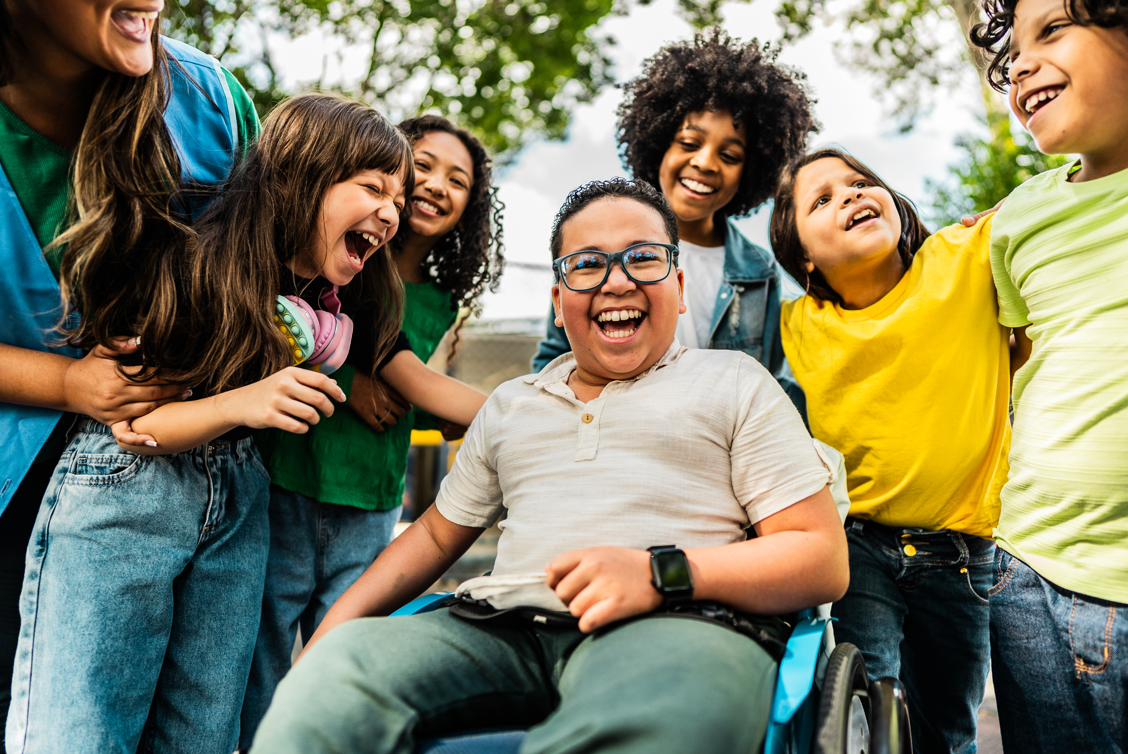 Smiling children gathered around a boy in a wheelchair outdoors.
