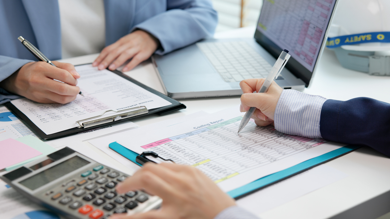 Two people review financial documents with a calculator and laptop on a desk.