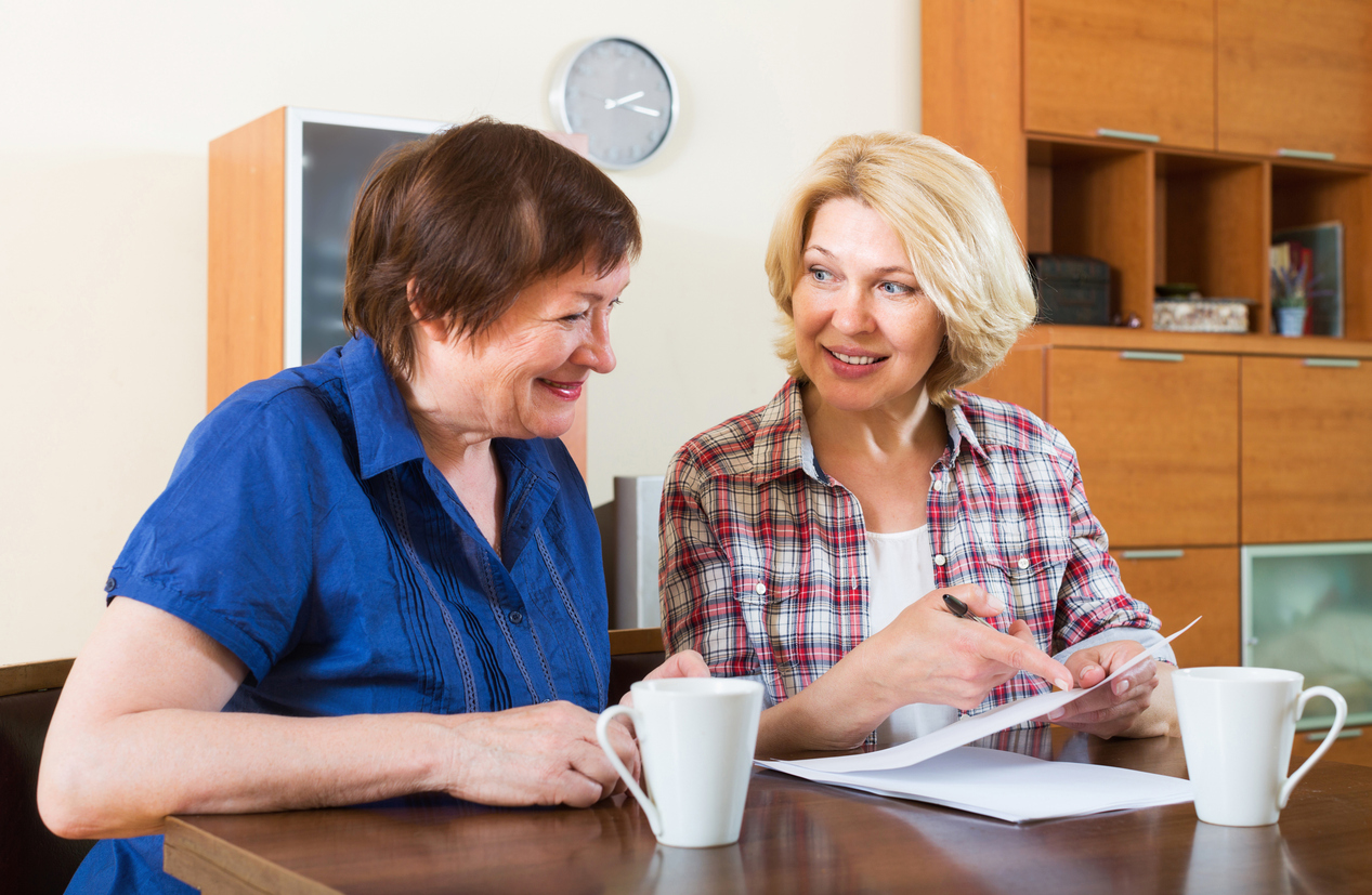 Two women smiling and discussing papers at a table with coffee mugs.