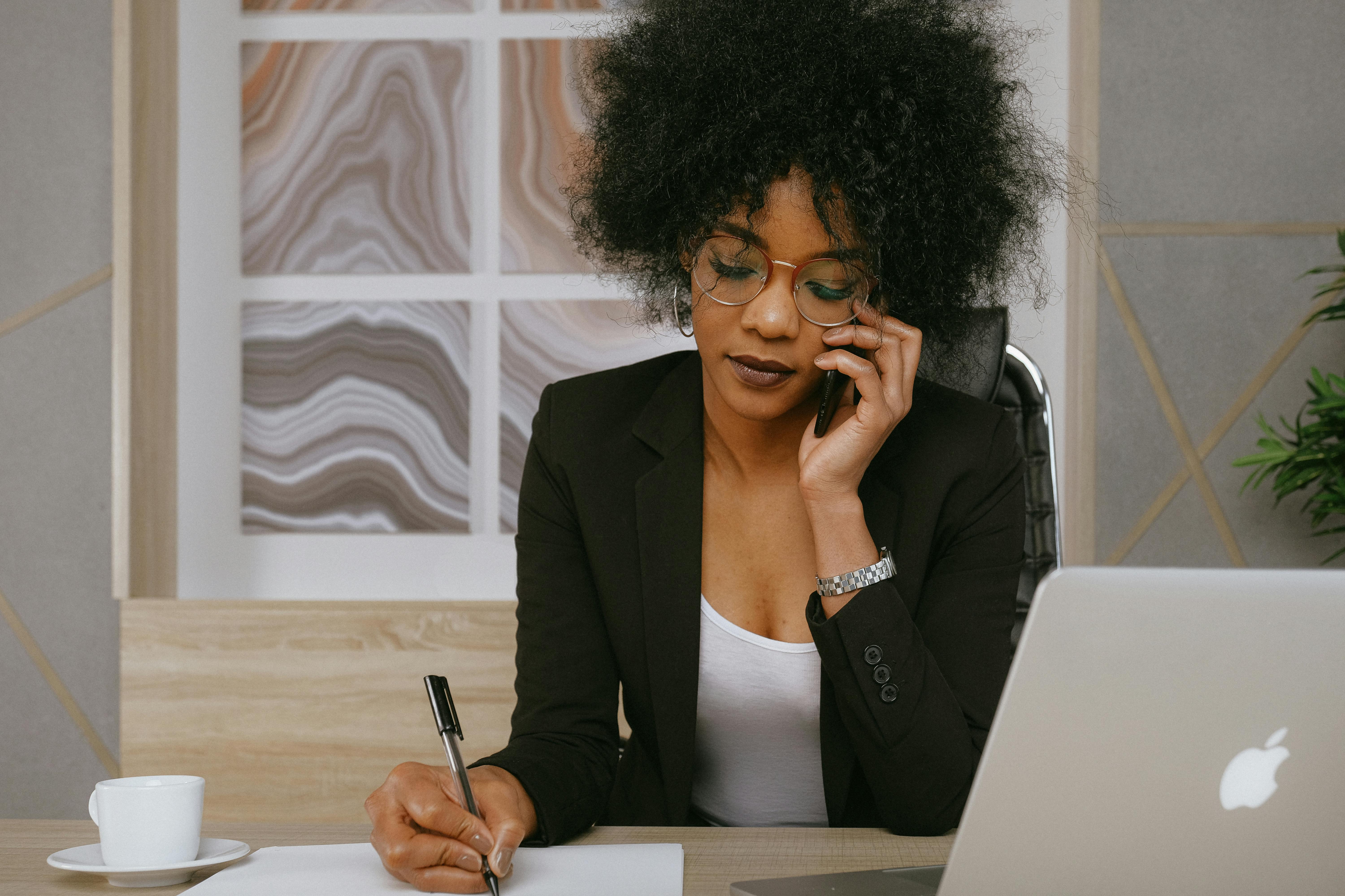 Woman in business attire talking on phone, writing notes at desk with laptop and coffee.