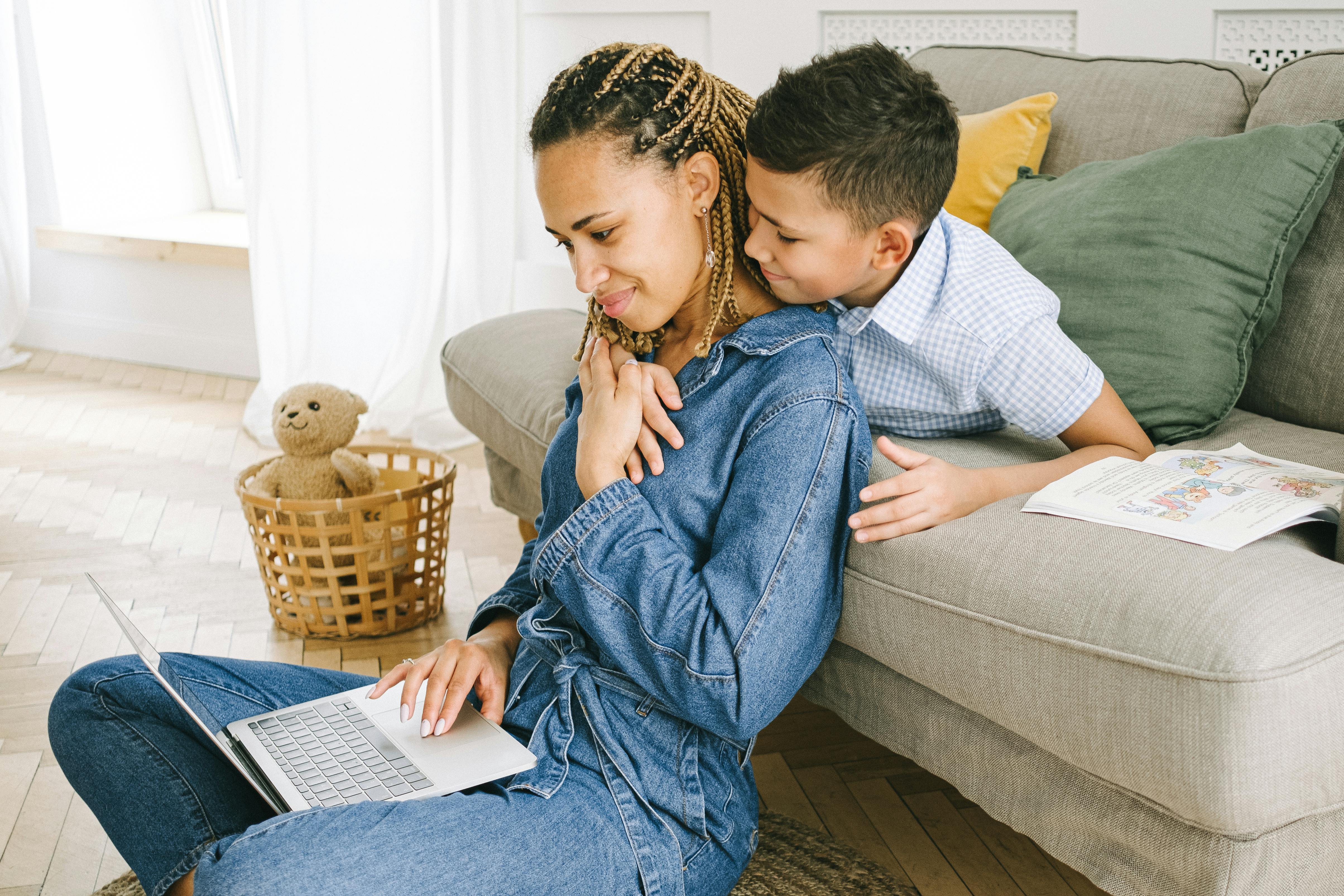 Smiling boy hugs woman working on laptop near a couch in a cozy living room.