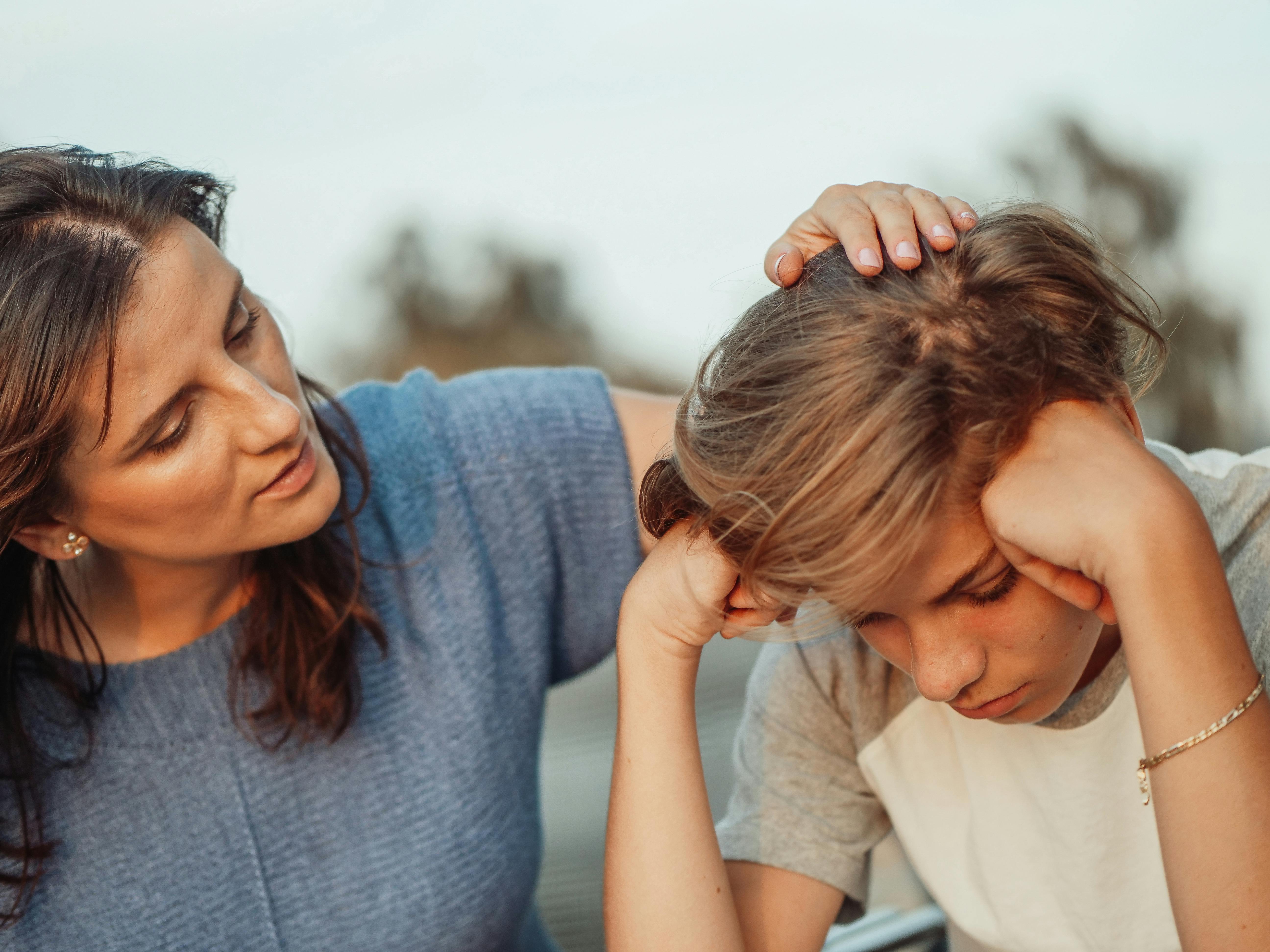 Woman comforting a sad child outdoors, hand gently on their head.