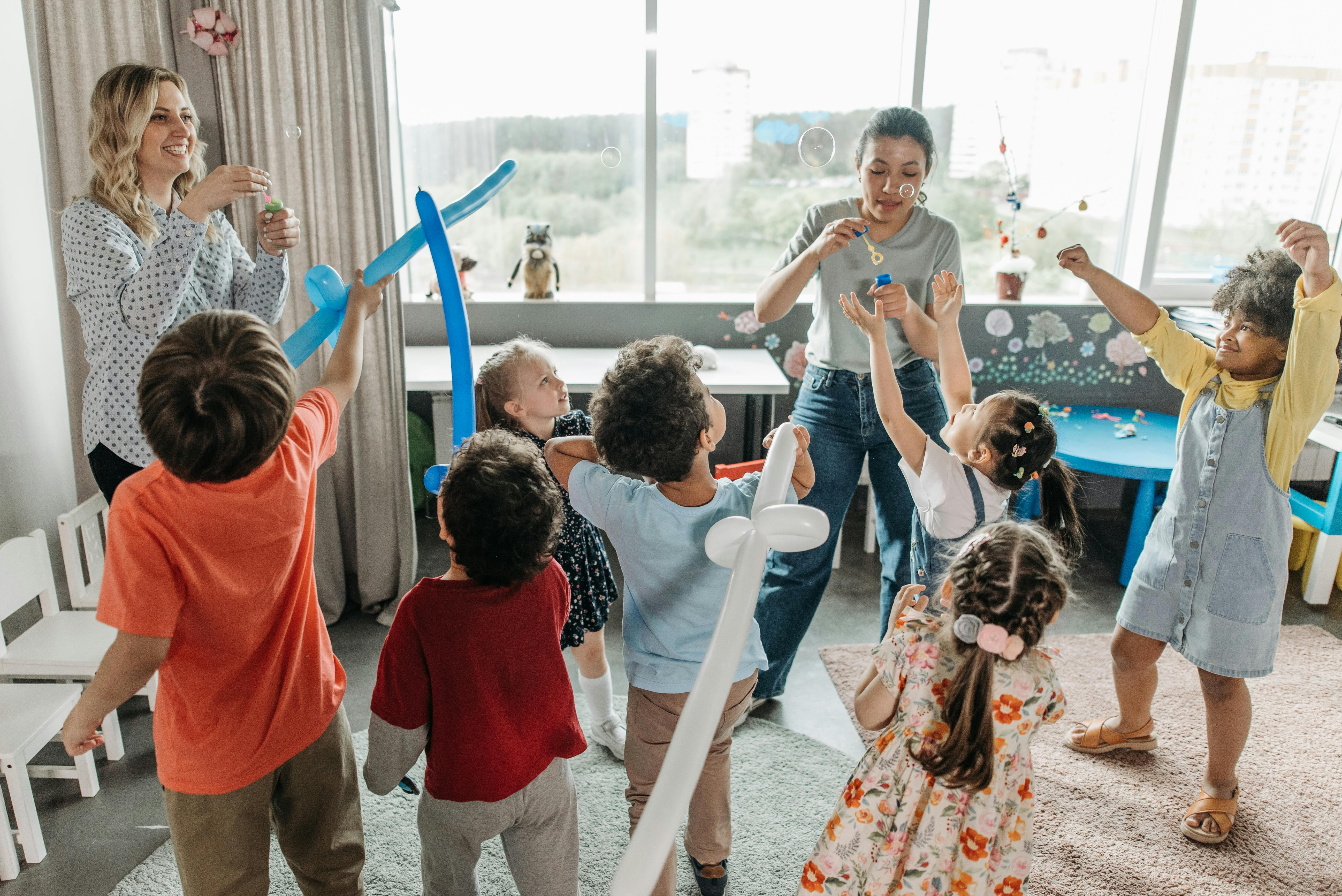 Children and two adults playing and laughing together in a brightly lit room blowing bubbles..
