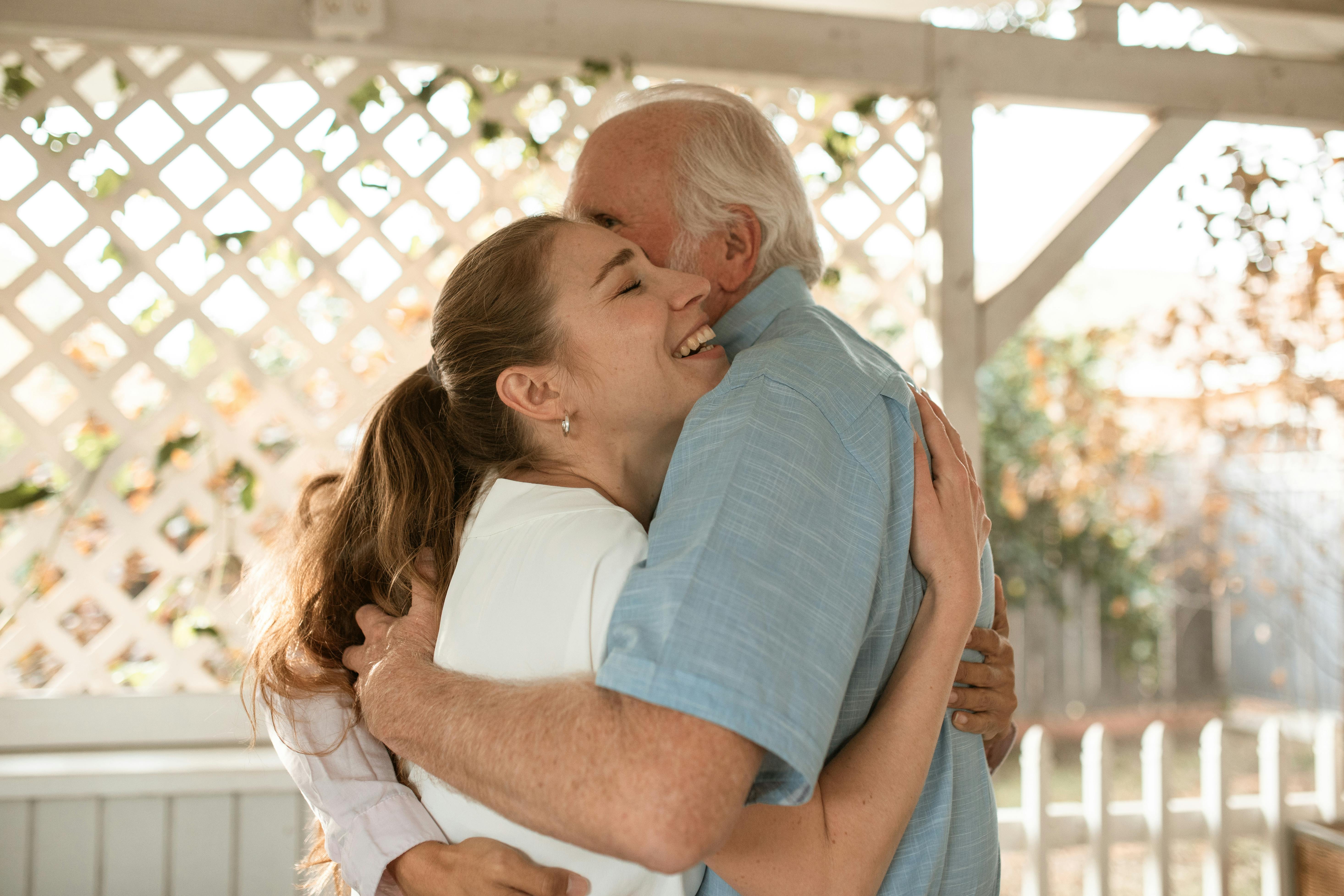 Young woman and older man smiling and hugging in a sunlit outdoor setting.