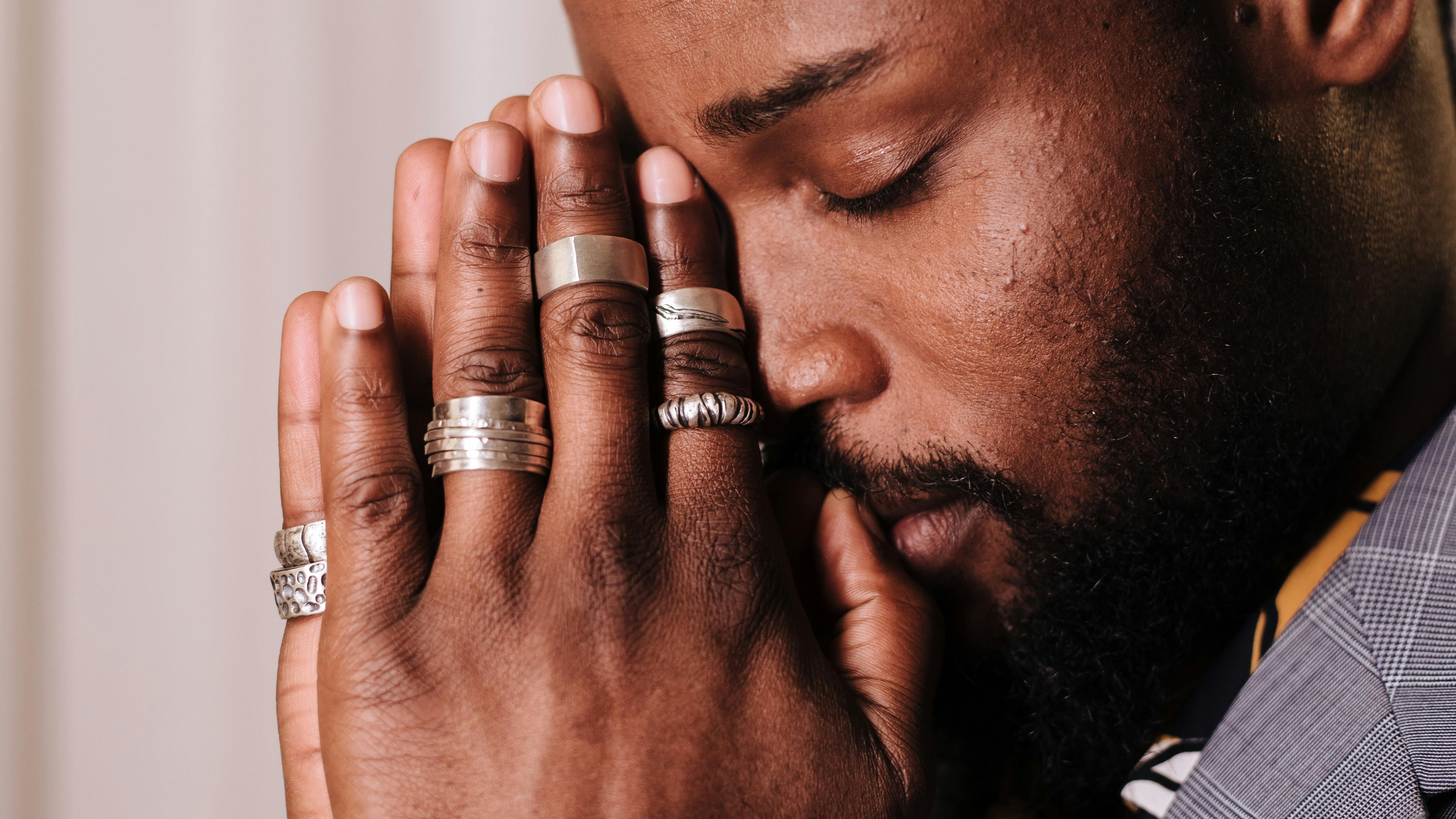 Man with closed eyes holding hands together to pray, wearing multiple rings.