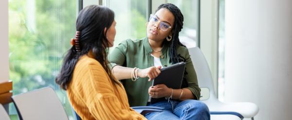 Two women sitting and talking by large windows in a bright, modern room.