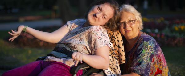 Woman in wheelchair smiling with older woman hugging her in a park.