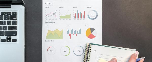 Overhead view of a work desk with laptop, printed analytics dashboard displaying various charts and graphs, and hand taking notes in a notebook.