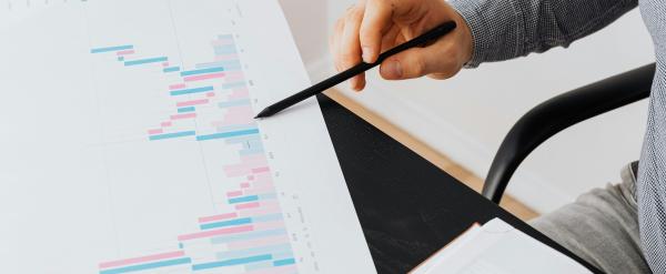 Hand pointing at a colorful Gantt chart on paper next to an open notebook.