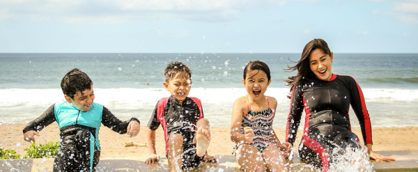 Three smiling children with Mom splash water at the beach on a sunny day.