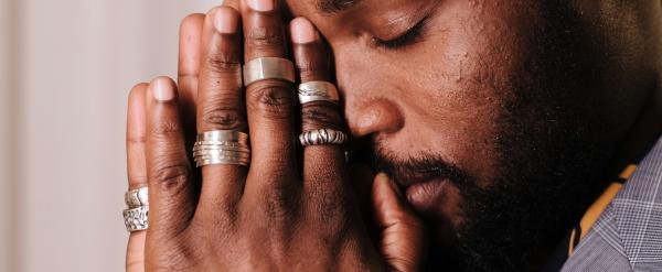 Man with closed eyes holding hands together to pray, wearing multiple rings.