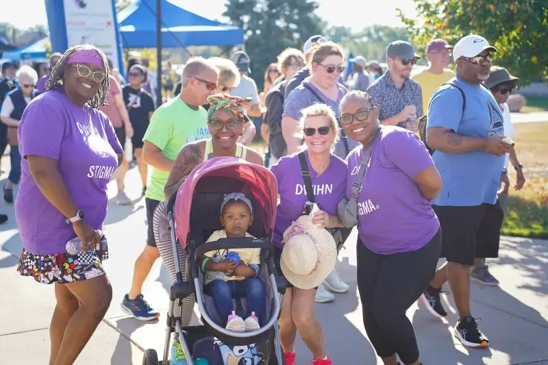 Group of smiling adults and a child in a stroller at an outdoor event, some wearing purple shirts.