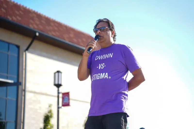 Man in a purple shirt speaking into a microphone outdoors on a sunny day.
