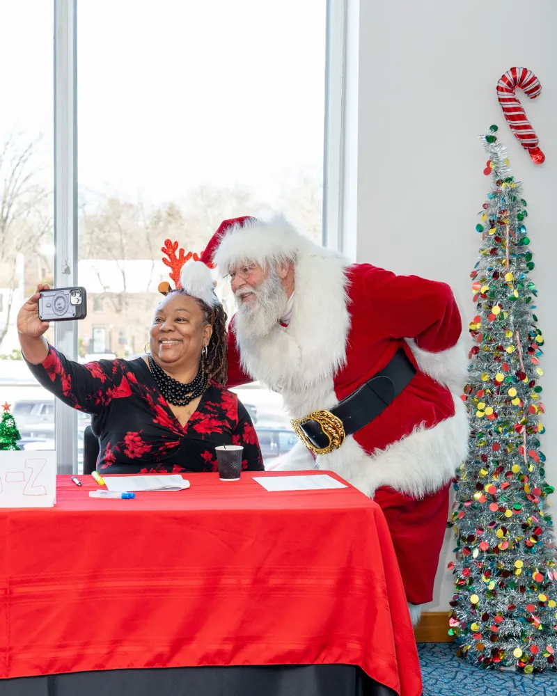 Woman in reindeer antlers and Santa take a selfie at a festive table near a decorated tree.