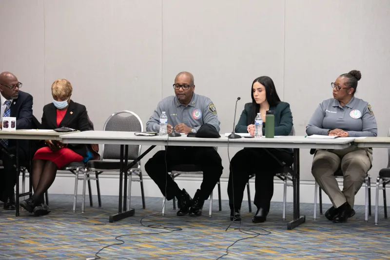 Five people sitting at a long table in a conference room, two wearing matching gray shirts.