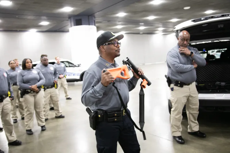 Officer leading a police training session indoors, holding a training weapon.
