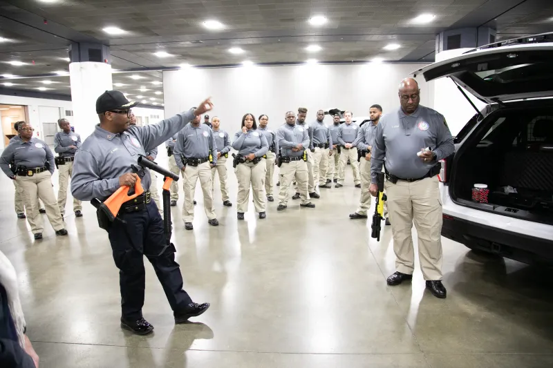 Security personnel in uniform stand in formation as one person gives instructions indoors.