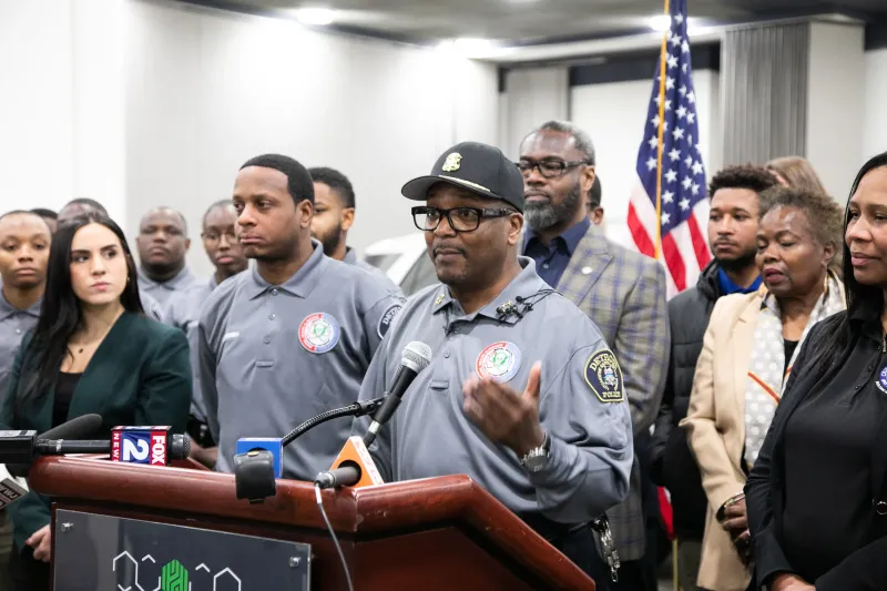 Man in uniform speaks at a podium surrounded by diverse group, US flag in background.