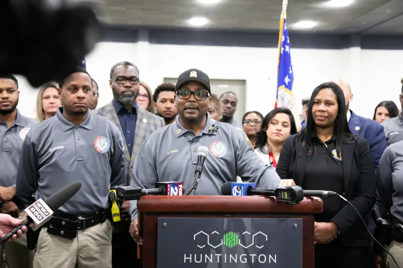 Man speaking at a podium surrounded by a diverse group, microphones and American flag visible.