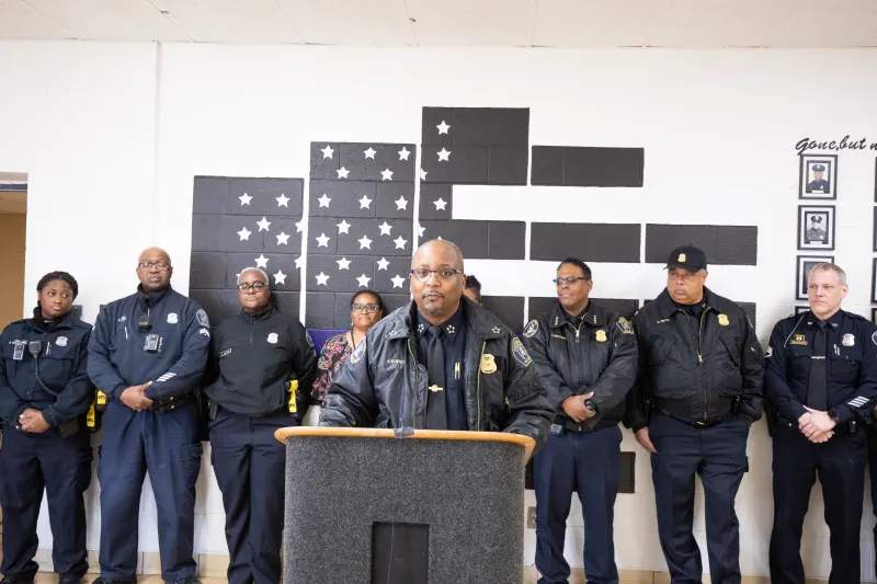 Police officer speaking at a podium with a group of officers standing behind him.