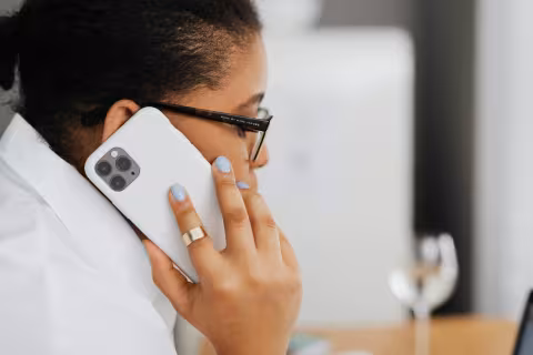 Person with glasses holding a smartphone to their ear, sitting at a desk.