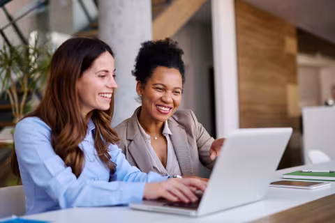 Two women smiling while working together at a laptop in a modern office.