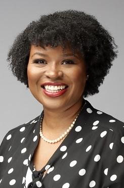 Smiling woman with curly hair, red lipstick, polka dot blouse, and pearl necklace.