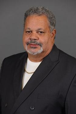 Man with gray hair and beard wearing a black suit and white shirt, neutral background.