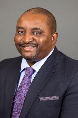 Man in a dark suit and purple tie smiling against a gray background.