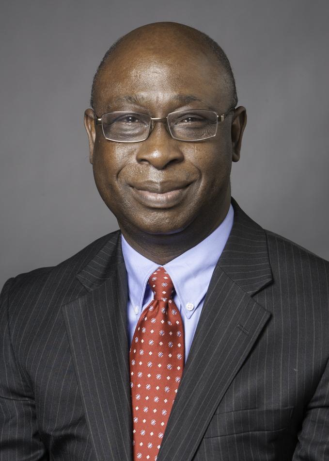 Man in a dark suit and red polka dot tie smiling against a gray background.
