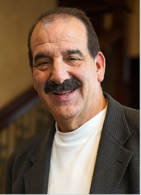 Smiling man with a mustache in a blazer and white shirt, indoors.