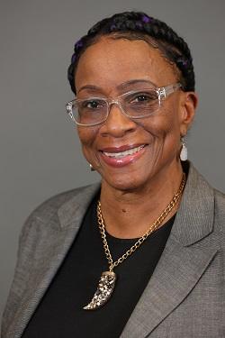 Smiling woman in gray blazer with braided hair and glasses on a gray background.