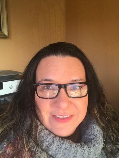 Woman with long dark hair and glasses smiling indoors, wearing a gray scarf.