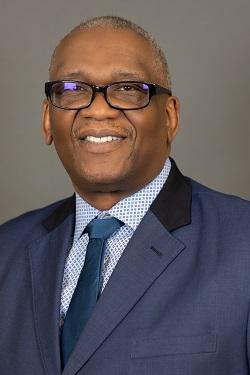 Smiling man in a blue suit, glasses, and tie against a gray background.